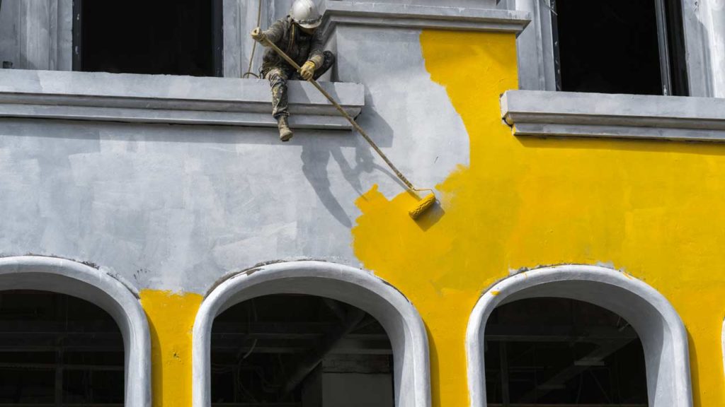 Man on ledge of building at a commercial painting job painting with an extended roller